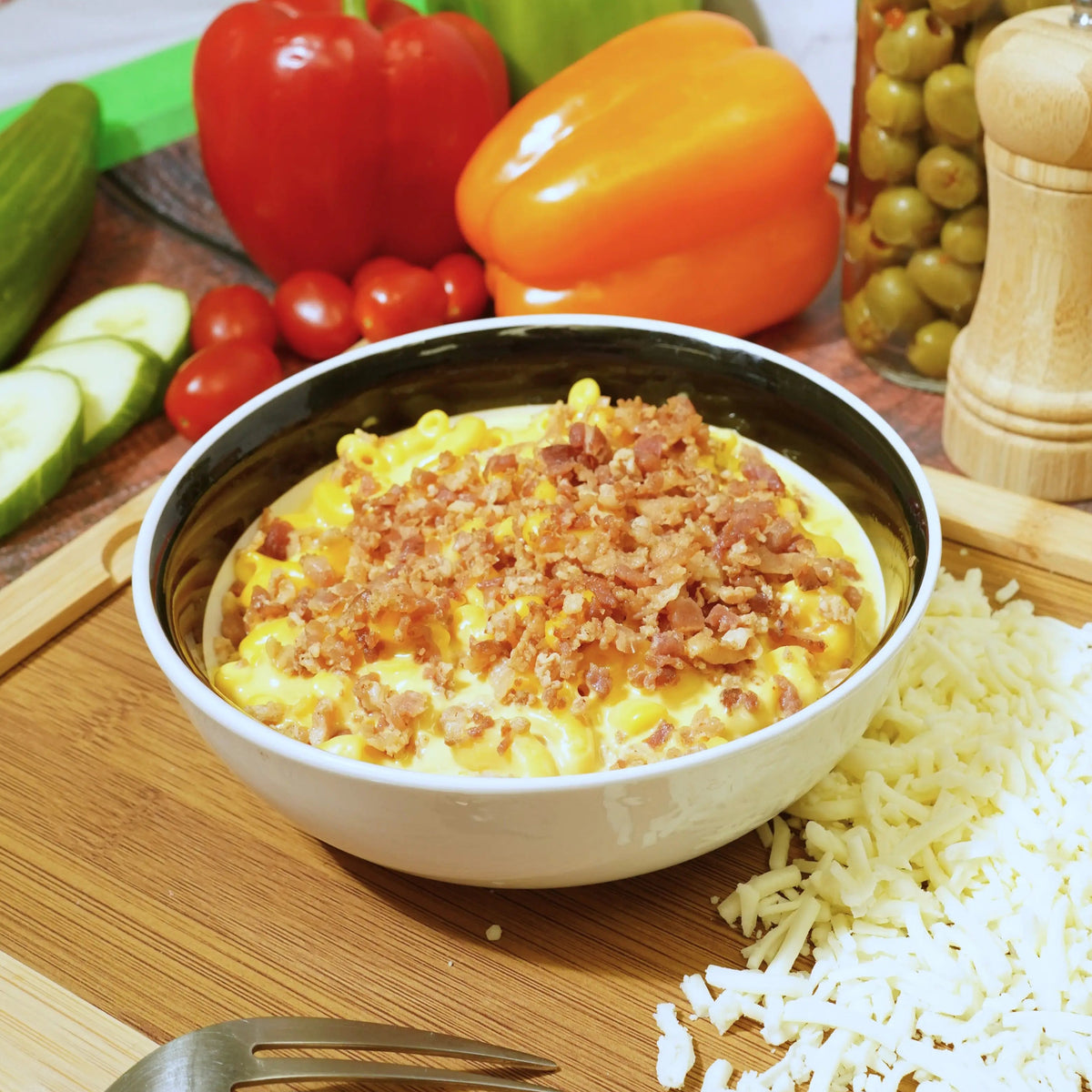 Bowl of mac and cheese with bacon on a wooden cutting board with vegetables and cheese.