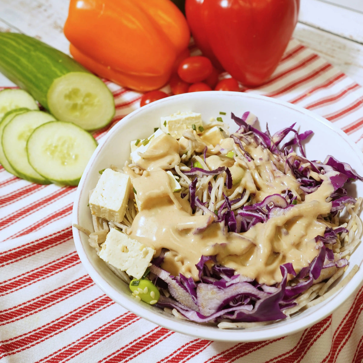Salad with tofu and vegetables on a striped tablecloth with bell peppers and cucumbers.