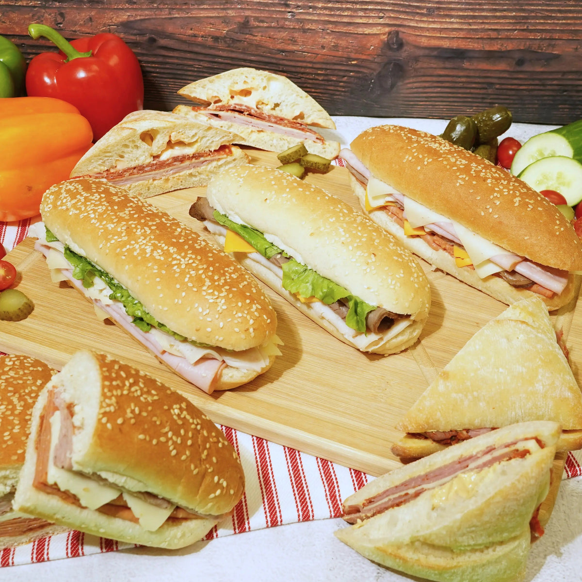 Assorted sandwiches on a wooden board with vegetables and chips on a checkered cloth.